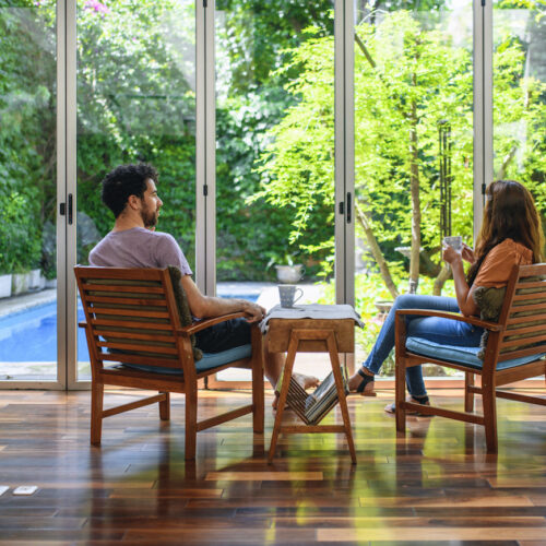 Rear view of Hispanic female and mixed race male in 20s and 30s sitting with coffee in naturally lit home sitting area with view of backyard and pool.